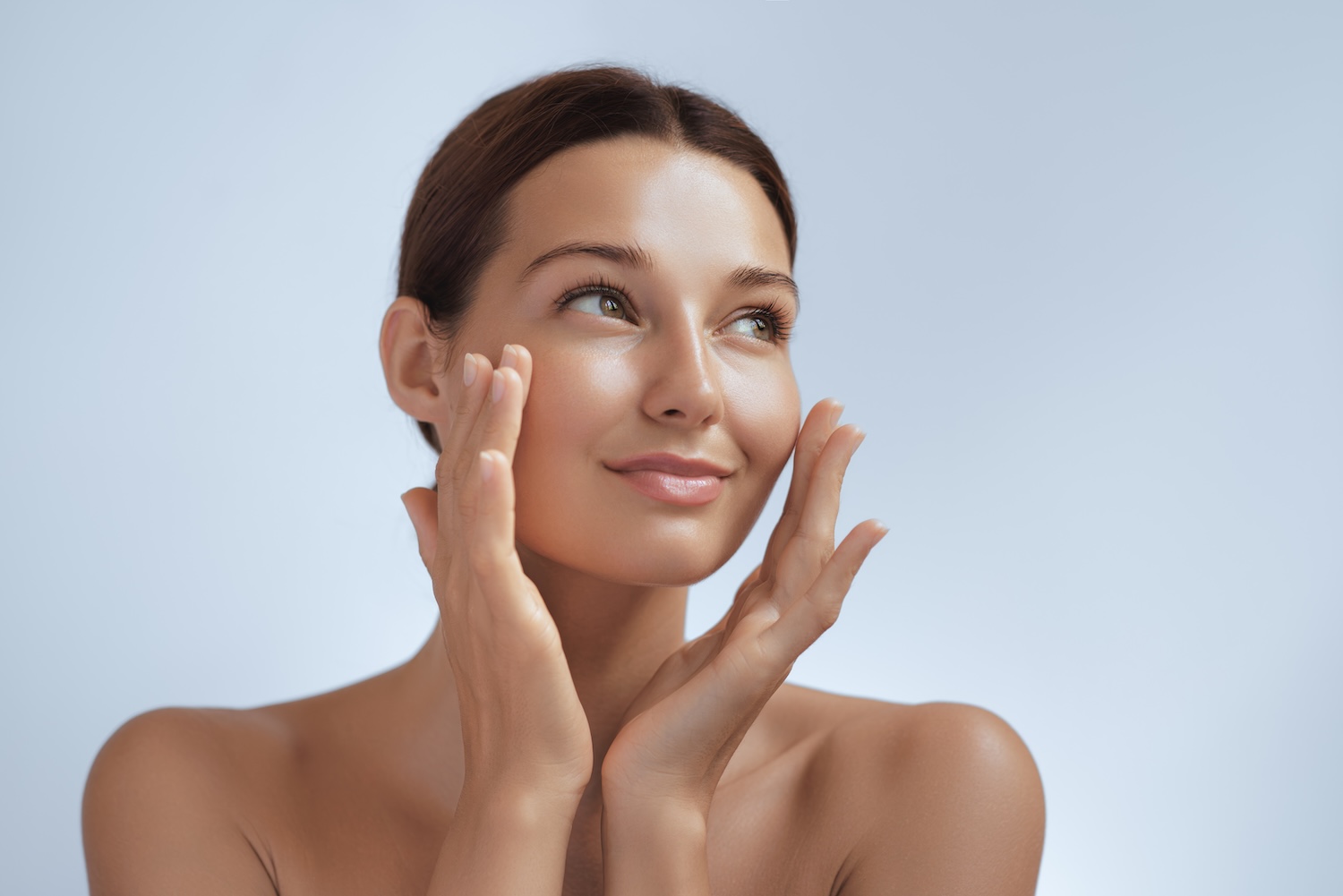 woman looking upward with hands gently touching her cheeks against a light background