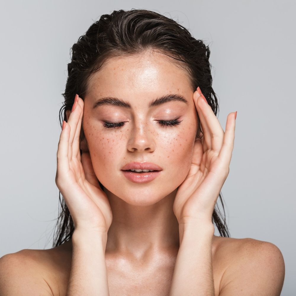 Woman with glowing freckled skin holding both hands to her face with eyes closed against a neutral background